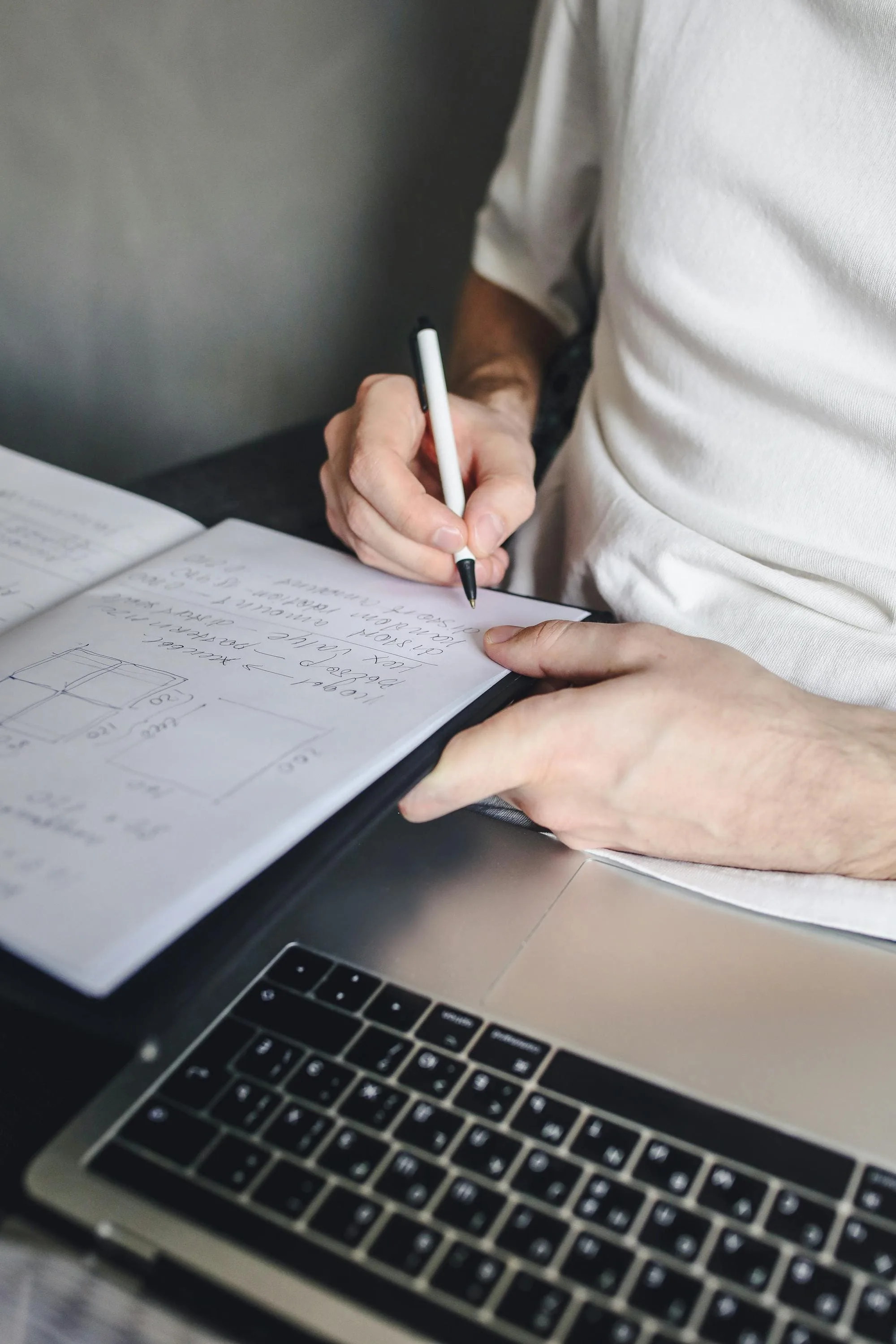 Person writing notes in a notebook next to a laptop